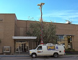 In front of a building entrance, a white truck with a raised large antenna. On the side of the truck, as well as on the driver's side door, are large "FOX 8 WJW CLEVELAND" signs.