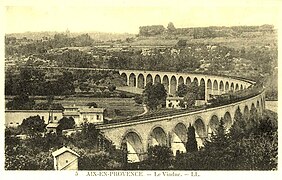 Photographie en sépia montrant un train composé d'une locomotive coupe-vent de de plusieurs voitures sur le pont, il décrit une trajectoire en courbe et la campagne autour du viaduc est peu urbanisée.