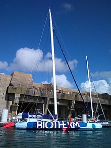 Vu de profil, en entier, bateau en camaïeu de bleu, amarré au ponton devant un gigantesque bunker.