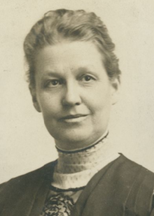 A middle-aged white woman, slightly smiling, wearing a high-collared lace-trimmed blouse under an academic robe