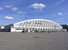 Exterior of hockey arena with an arched roof clad in white-colored siding, with multiple flags on poles in the foreground