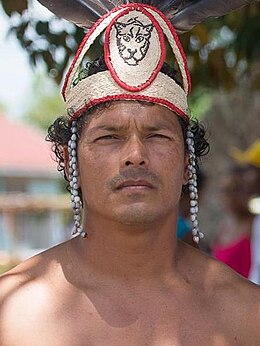 Portrait photographique en plan buste et en couleur. Un homme portant une coiffe amérindienne se tient en extérieur, les épaules nues. Flou d’arrière-plan laissant transparaître un bâtiment et de la végétation. Temps ensoleillé.