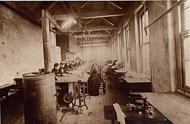 Photographie en nuances de sépia. Dans une grande salle tout en longueur aux combles apparentes sous un toit pentu, des femmes en robes sombres et en chignons sont assises sur des tabourets à des postes de travail pourvus de machines à coudre mécaniques. La pièce est éclairée par des baies vitrées sur la droite. À gauche au premier plan, un grand poêle en fonte chauffe la salle.