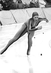 A woman is wearing a skin-tight jumpsuit with a hood and ice skates; she is making a turn as she skates on an ice track.