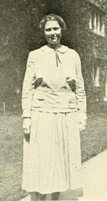 A young white woman standing outdoors, wearing a light-colored dress with peter-pan collar and long sleeves