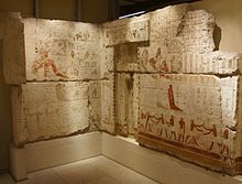 View of two limestone walls (composed of larger blocks) at right angles to each other, with coloured reliefs in dark red, ochre and blue. A false door has been inserted near the corner of the walls.