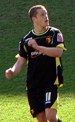 A man wearing a predominantly black shirt and shorts, both with yellow and white patterns, standing on a grass field.