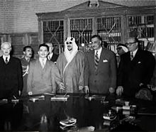 Five men standing side by side behind a table with documents on it. All the men are wearing suits and ties, with the exception of the man in the middle, who is wearing a traditional robe and headdress. There are three men standing behind them.