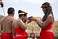 Deux femmes, un homme, vêtus de sortes de pagnes rouges. Une femme boit dans un grand bol.