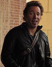 A Caucasian male with brown hair smiling while raising his right eyebrow. He is wearing an unzipped black leather jacket over a dark colored shirt. In the background, a yellow and red bricked wall can be seen