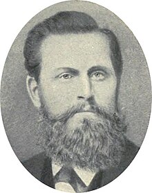 Photograph of man in a suit and tie with dark hair and a bushy beard