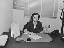 Photograph of a woman with short hair wearing a dark-colored v-necked dress, sitting at a desk in an office