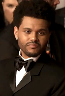 Picture of the Weeknd (Tesfaye). He is wearing a black suit with a bowtie, averts his gaze from the camera, and has a neutral expression on his face.