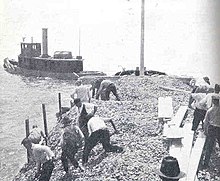 men shoveling shells into water from scow being towed by another boat