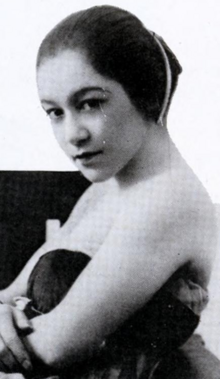 A young woman with dark hair dressed back, away from the face, gathered in a smooth coiffure at the nape. She is wearing a strapless dress.