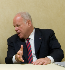 Photo of a man with grey hair and a black suit sitting on a table and talking to someone on his right