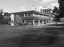 black and white image of a modernist building, two stories at the front, four stories at the back with trees and roads around it