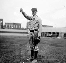 A black-and-white photograph of a man wearing an old-style baseball uniform and a newsie cap holding a baseball in his outstretched hand