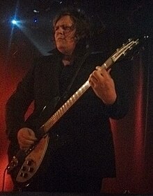 Rover wearing a dark suit, standing onstage holding a guitar, looking intently right of camera