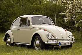 A front-three quarters view of a pale-yellow Volkswagen Käfer. It features 165/80R15 tires, which shod 15x4. 5" silver, circular wheels. The Käfer features a beetle-like body, and its window is open. The picture is taken with much greenery in the background, and the photo was edited to give it a more warmer tone.