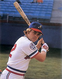 A man wearing a white baseball uniform with a blue cap poses holding a baseball bat with both hands posed as if ready to swing