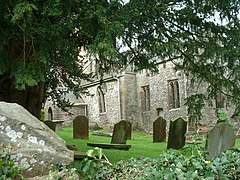 Side of stone building with arched windows, partially obscured by trees. Gravestones in the foreground.