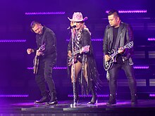 Gaga in a pink cowboy hat plays guitar on a purple-lit stage, flanked by two other guitarists.