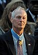 Head and shoulders of an older white man with a large scar above his left eyebrow. He is wearing a dark suit coat with an American flag pin on the lapel and a star-shaped medal hangs from a light blue ribbon around his neck.