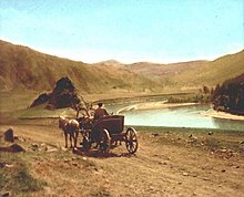 Calèche avec un paysan et deux chevaux longeant une rivière, présumément la Tchouïa, au début du XXe siècle. La scène est représentée sur une carte postale. En arrière-plan se trouve un paysage montagneux.