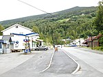 Foto einer Straße, die durch eine Ortschaft führt. An der Straße liegt unter anderem eine Tankstelle sowie mehrere weiße Holzhäuser.