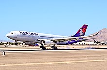 A white twin-engine plane painted with the word "HAWAIIAN" in the front and a woman in different purple hues on the tail has just landed on a runway on a sunny day amid a desert backdrop