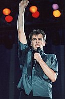 Peter Gabriel wearing a blue shirt, singing into a microphone while holding one fist in the air