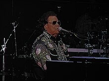 Ronnie Milsap in a sequined black suit and sunglasses, seated at a piano while singing into a microphone