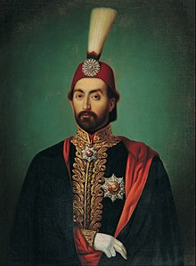 Portrait du sultan ottoman Abdülmecid Ier à la barbe courte portant un fez surmonté d'un plumeau, un manteau noir doublé de rouge et une veste aux broderies dorées sur laquelle se trouvent plusieurs médailles en forme d'étoiles.