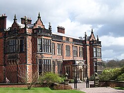 A brick hall seen from a slight angle with bay windows, brick diapering, a porch, shaped gables, and statues standing on the parapets