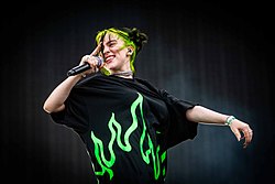 Image of a woman singing into a microphone. She sports green-black hair and clothing and stands against a black backdrop.