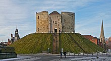 A photograph of a small castle on top of a green mound; the castle has three circular walls visible. Behind the castle the sky is overcast and dark grey.