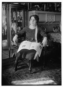 A young woman with fair skin and dark hair, seated indoors, with a magazine or newspaper open on her lap