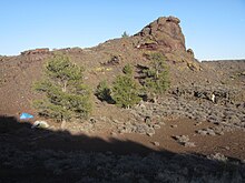 two tents and a person next to the wall of an open crater lined with some sagebrush and four pine trees