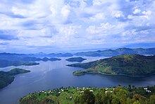 Der See weist einige unterschiedlich große Inseln und rundum Hügel auf. Der Himmel ist stark bewölkt.