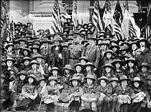 A crowd of white girls in Girl Scout uniforms, surrounding an older white woman, a white man in uniforrm, and another white woman in uniform; photographed outdoors, in front of an array of flags