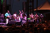 All-male group on stage containing a guitarist, a drummer, a vocalist, a keyboardist, and three members of a horn section. Some of the crowd present in the foreground