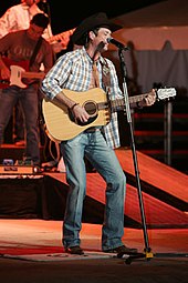 A man wearing a black cowboy hat, a checked shirt and jeans, playing guitar and singing into a microphone