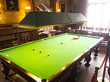 A full-size snooker table in a brightly lit room with bookcases and a boardroom table in the background, all cordoned off at the right-hand side as part of an English country house display