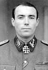 A man wearing a military uniform and neck order in the shape of a cross. His black hair is parted and his facial expression is determined.