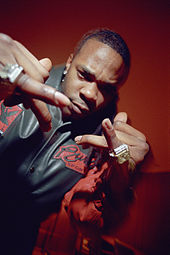 A picture of a man displaying the jewelry on his fingers to the camera. He wears a red-black jacket and stands in front of a red background.