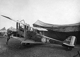 Single-engined military biplane with two men in the cockpit, parked in front of a large tent