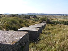 Area of grassy sand dunes with a row of large concrete cubes across it. People at a bridge in the distance.