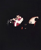 A bearded man wearing a bowler hat and waistcoat playing a clarinet under a spotlight