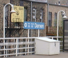 A blue rectangular sign saying "Damen" in the center surrounding by ancillary information is flanked on either side by two light fixtures that protrude from the railing and curve down back towards the platform, providing the so-called "gooseneck" shape. Both the railings and the light fixtures are painted white.